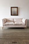 A two-seater sofa upholstered in a beige and white vertically striped fabric, with light wood legs, set against a white wall with a framed mirror above and a wooden floor below.