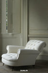 A white leaning club chair with buttons, featuring a turned front leg and a square backrest, placed in a room with visible wall panels and a window.