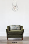 Green armchair with a patterned cushion under a geometric pendant light on a white wall.