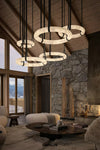 A luxury modern chandelier with multiple interlocking circular ring lights in a rustic mountain chalet living room. The brand name 'DUNLIN' is visible at the bottom.