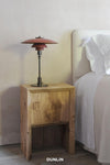 A rustic wooden bedside table next to a bed with white bedding, featuring a red Pagodas-style table lamp on top. The brand name 'DUNLIN' is printed in white text at the bottom.