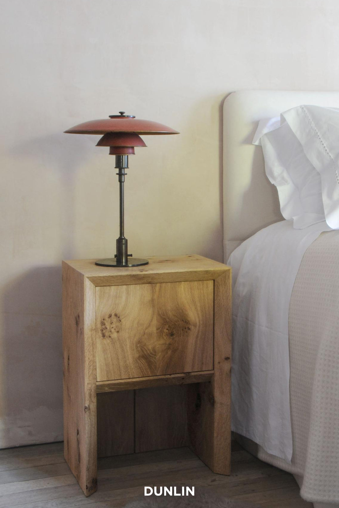 A rustic wooden bedside table next to a bed with white bedding, featuring a red Pagodas-style table lamp on top. The brand name 'DUNLIN' is printed in white text at the bottom.