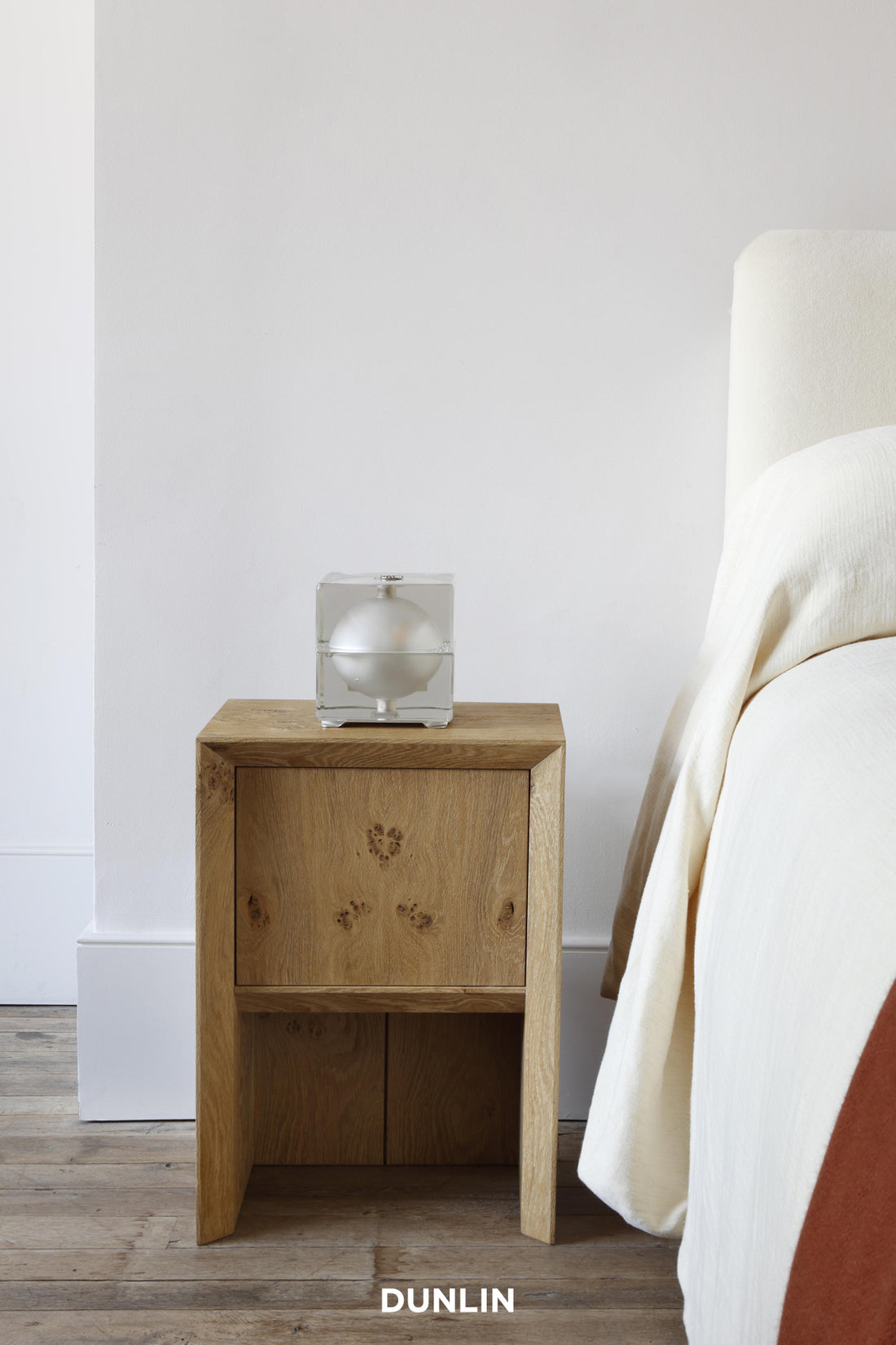 A light oak wood nightstand with a single drawer and carved floral details, styled next to a bed with cream bedding. A clear glass box containing a white orb sits on top.