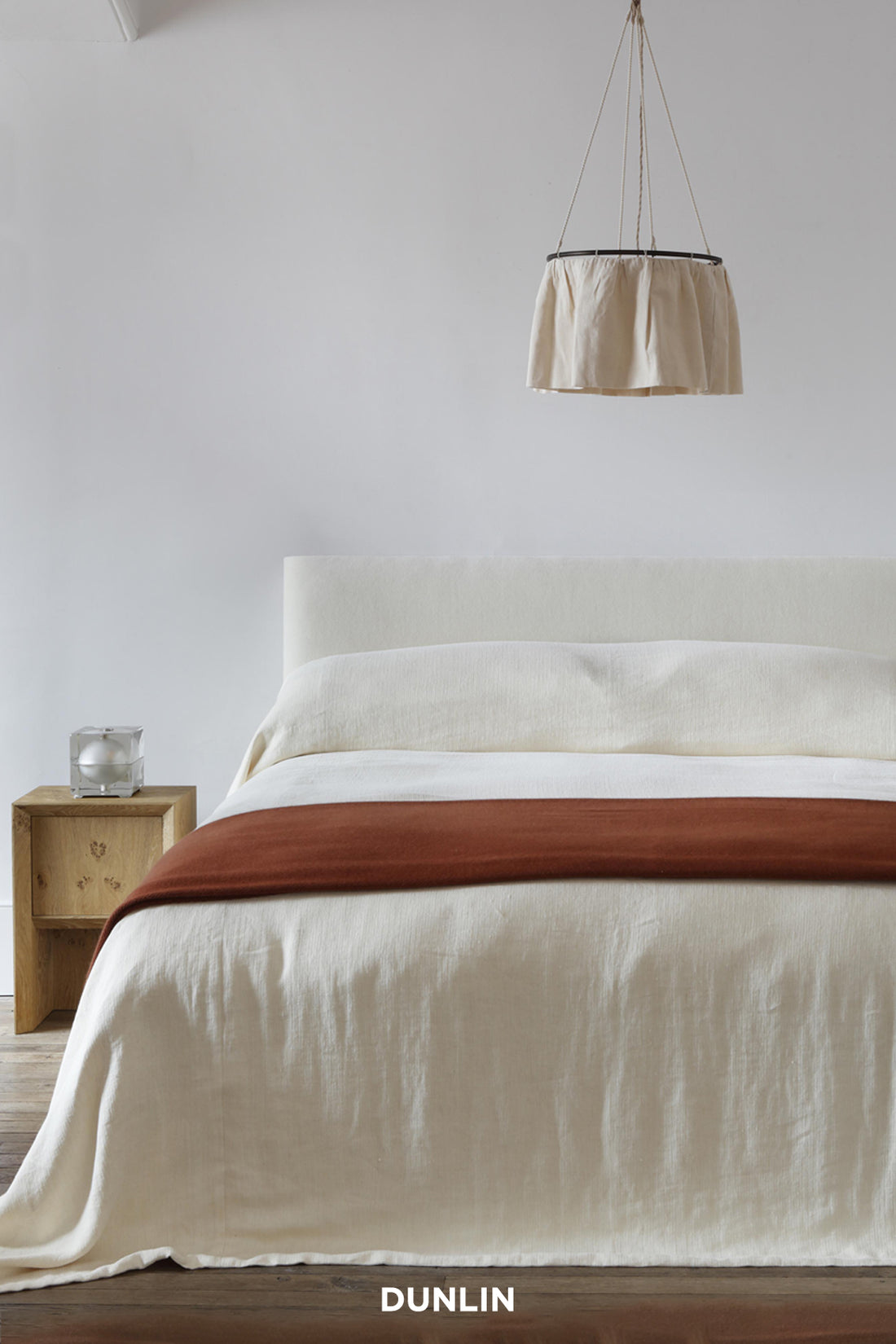 A minimalist bedroom scene featuring a bed dressed in off-white linen bedding with a rust-brown throw blanket across the foot. A light wood bedside table with a glass vessel sits to the left, and a beige fabric drum pendant light hangs from the ceiling above.