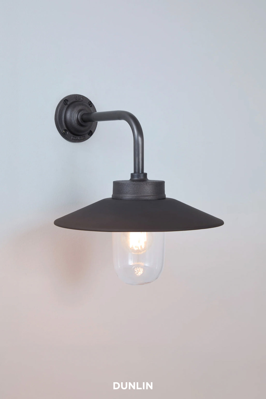 A wall-mounted industrial-style sconce light fixture in a dark gunmetal finish, featuring a wide conical shade, curved arm, and a glowing clear glass bulb. The brand name 'DUNLIN' is printed in white at the bottom.