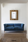 A blue velvet two-seater sofa with wooden legs, positioned in a room with a white wall, an ornate gold-framed mirror, and a wooden floor. The brand name 'DUNLIN' is printed at the bottom.