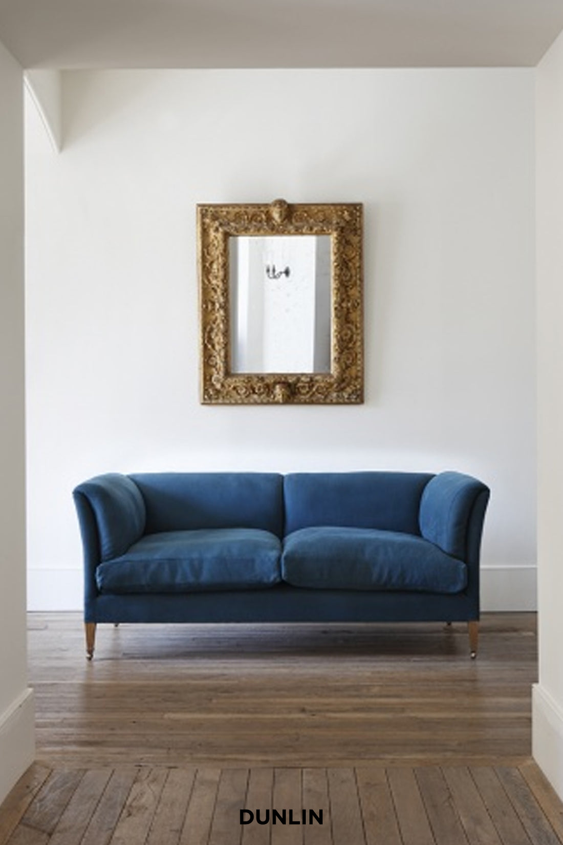 A blue velvet two-seater sofa with wooden legs, positioned in a room with a white wall, an ornate gold-framed mirror, and a wooden floor. The brand name 'DUNLIN' is printed at the bottom.