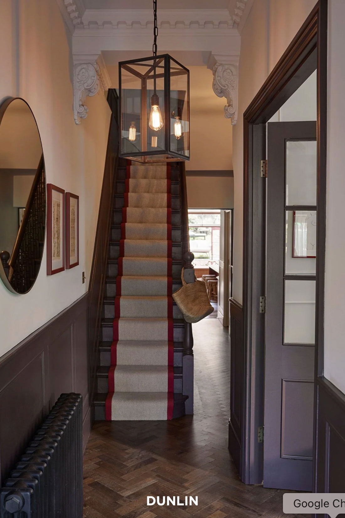 An interior hallway with a staircase featuring a beige carpet runner with red side borders, a hanging lantern-style pendant light, dark wood flooring, and a dark door on the right.