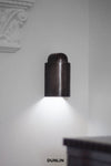 DUNLIN Decade Wall Light in Weathered Brass, featuring a cylindrical brass design with downward architectural illumination against a white wall.