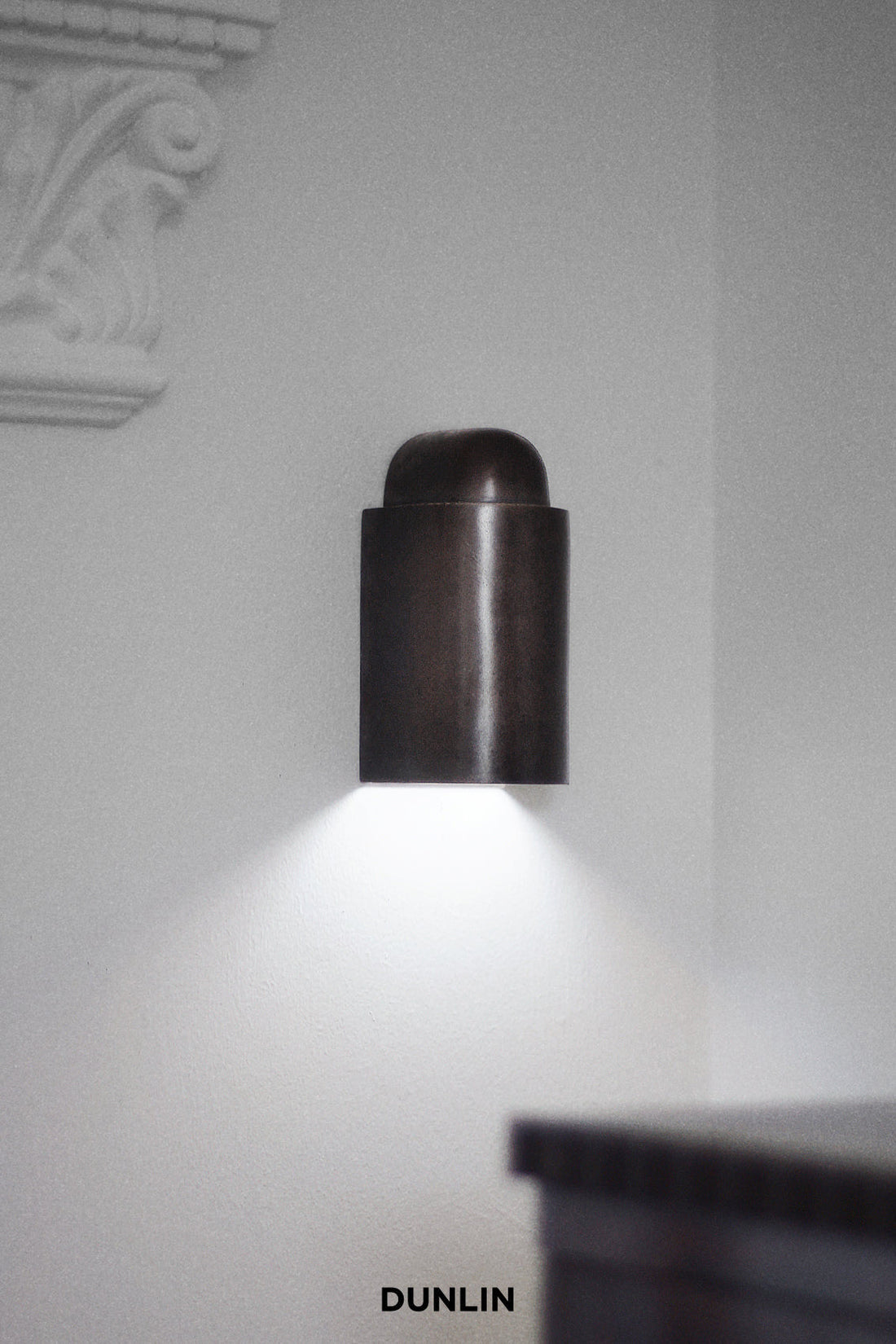 DUNLIN Decade Wall Light in Weathered Brass, featuring a cylindrical brass design with downward architectural illumination against a white wall.