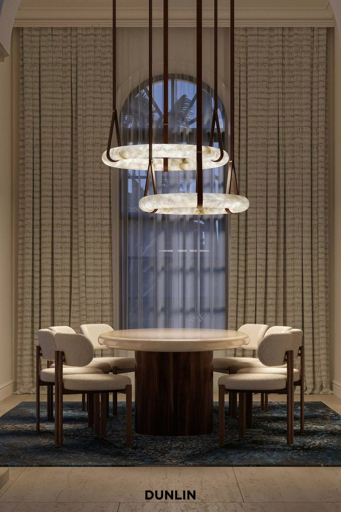 A luxury dining room setup featuring a round wooden dining table surrounded by four upholstered chairs, under a multi-tiered modern chandelier. The room is decorated with textured beige curtains, a blue patterned rug, and a large arched window in the background.