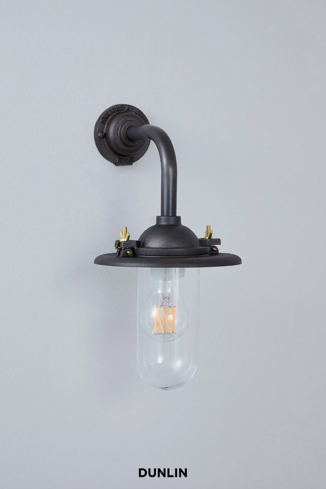 A wall-mounted industrial-style sconce with a dark gunmetal finish, featuring a curved pipe arm, a clear glass shade, and brass hardware, shown against a light gray wall with the brand name 'DUNLIN' printed below.
