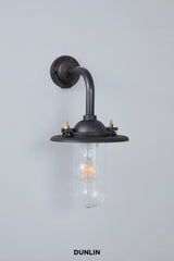 A wall-mounted industrial-style sconce with a dark gunmetal finish, featuring a curved pipe arm, a clear glass shade, and brass hardware, shown against a light gray wall with the brand name 'DUNLIN' printed below.