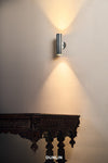 A nickel-plated brass up-down light fixture mounted on a wall, with illumination visible in the vertical and horizontal directions.