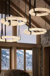 Four circular ring pendant lights with a textured cream-white finish and black hanging straps, suspended from a wooden ceiling in a rustic mountain chalet interior with snowy windows.