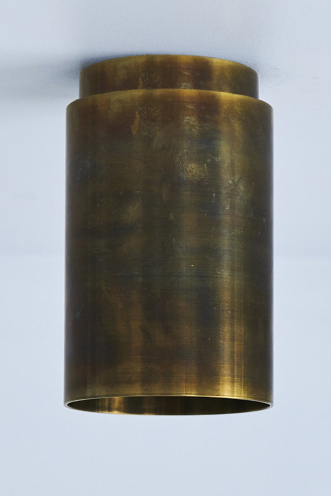 A cylindrical tarnished raw brass ceiling spotlight with a visible warm glow at the top.
