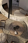 A top-down view of a modern living room featuring a large organic-shaped white marble console table, a round rustic wood coffee table, a black and white patterned rug, and light gray upholstered seating.