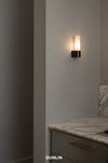 A modern wall-mounted sconce by Dunlin with a cylindrical frosted glass shade and a textured dark metal base, shown lit on a gray wall above a marble countertop. Palais Wall Light weathered Brass Glass LED Low Voltage DUNLIN