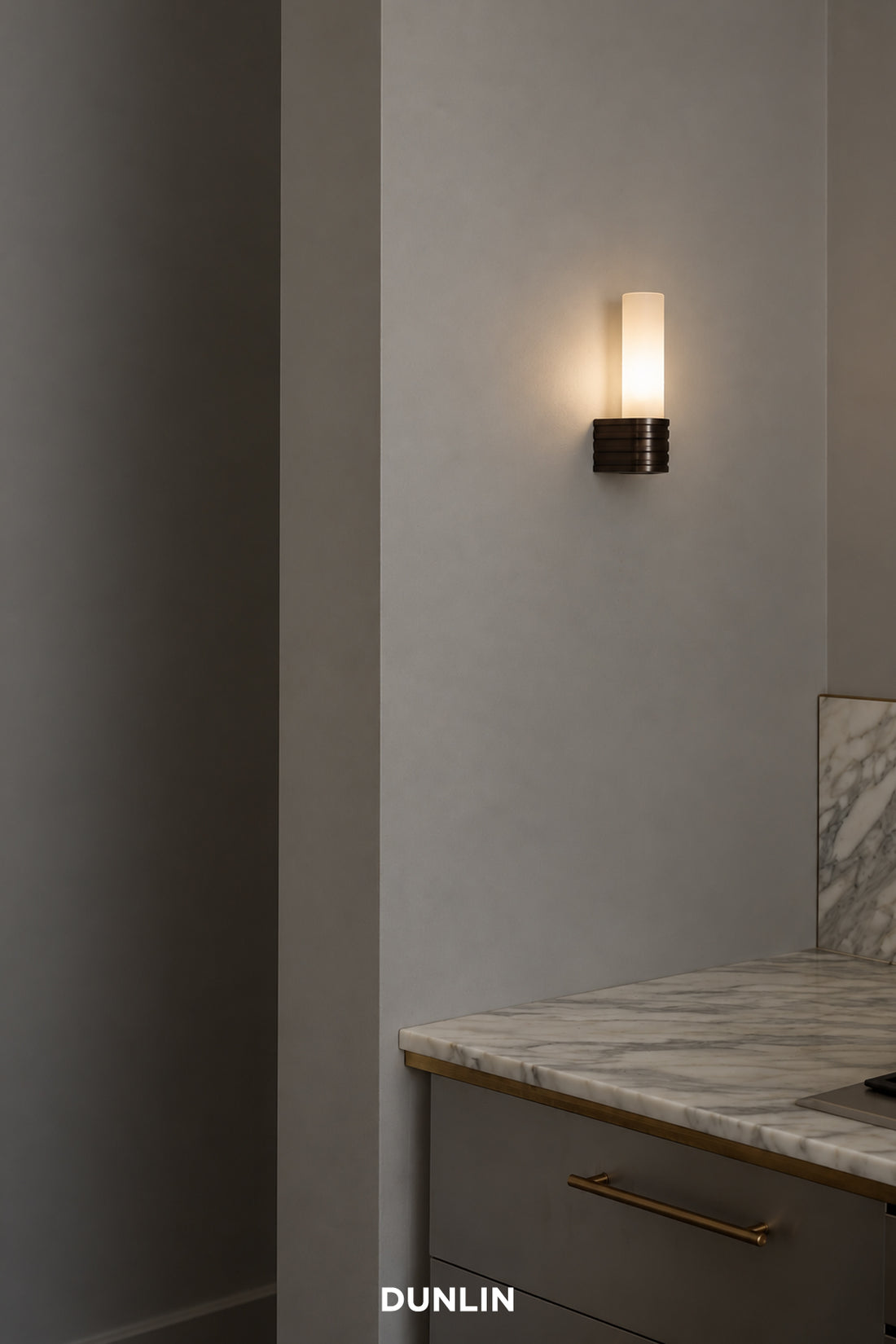 A modern wall-mounted sconce by Dunlin with a cylindrical frosted glass shade and a textured dark metal base, shown lit on a gray wall above a marble countertop. Palais Wall Light weathered Brass Glass LED Low Voltage DUNLIN