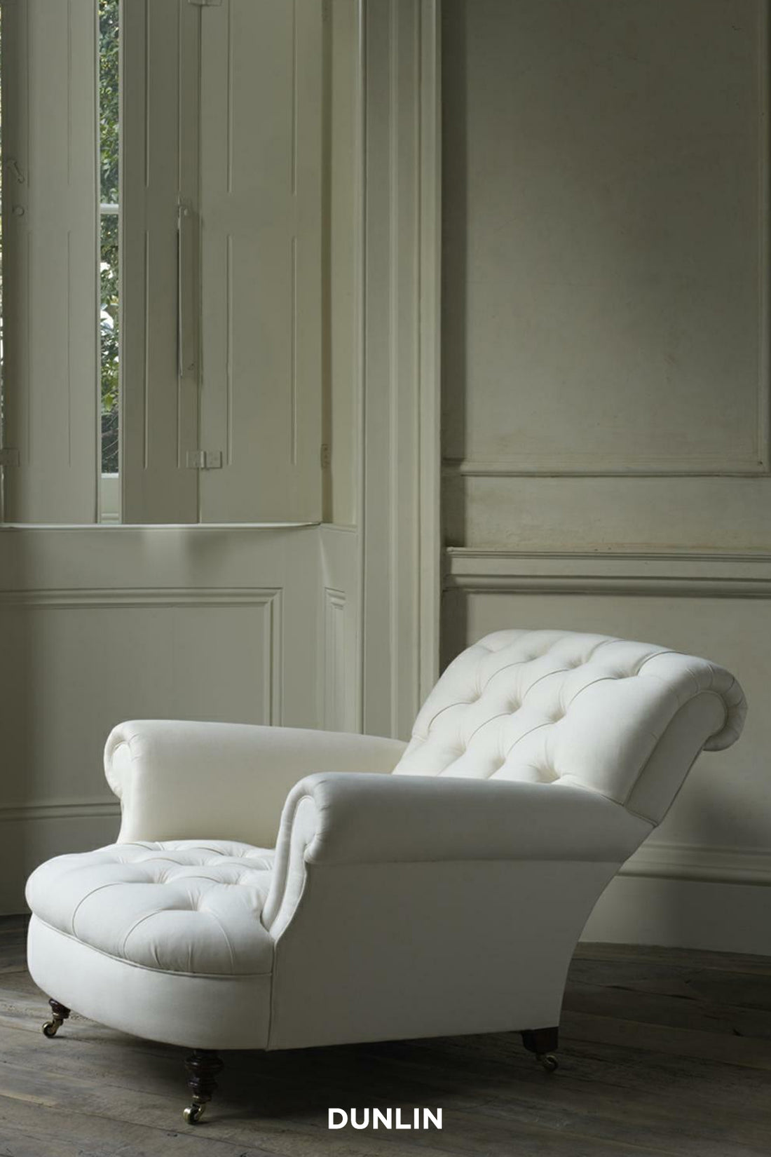 A white leaning club chair with buttons, featuring a turned front leg and a square backrest, placed in a room with visible wall panels and a window.