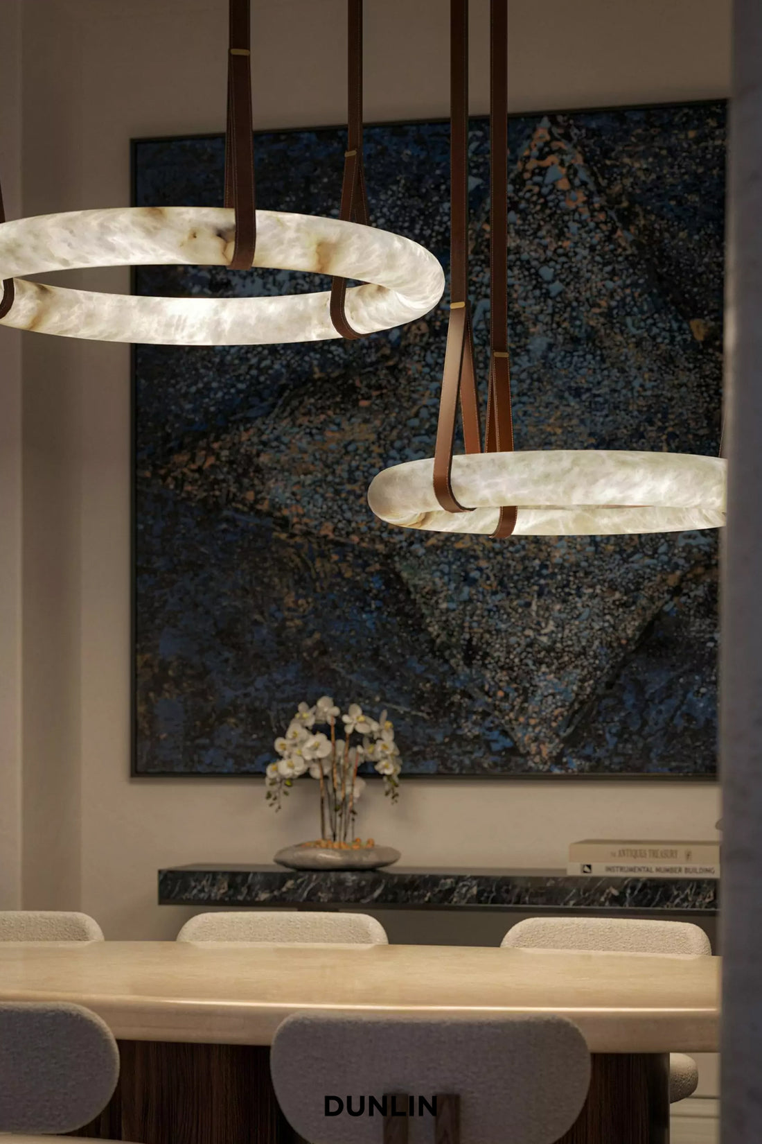 Two modern ring-shaped pendant lights with a milky white textured material and dark brown leather straps, hanging in a dining room setting above a table with chairs and a large abstract blue wall art piece.