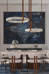 A modern dining room featuring a large abstract blue and gold wall art piece, with two circular pendant lights hanging above a long dining table and upholstered chairs.