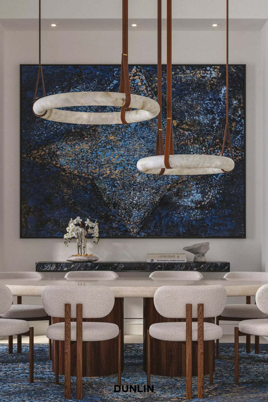A modern dining room featuring a large abstract blue and gold wall art piece, with two circular pendant lights hanging above a long dining table and upholstered chairs.
