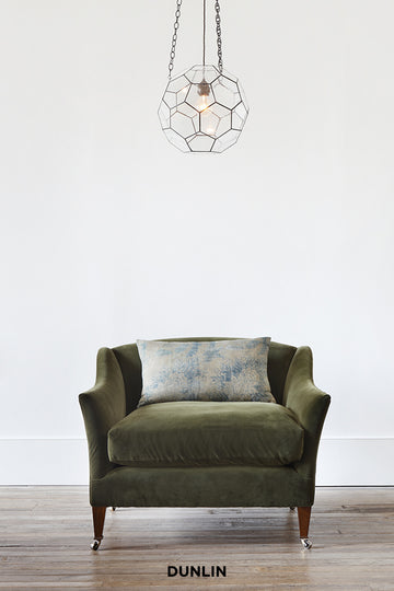 Green armchair with a patterned cushion under a geometric pendant light on a white wall.