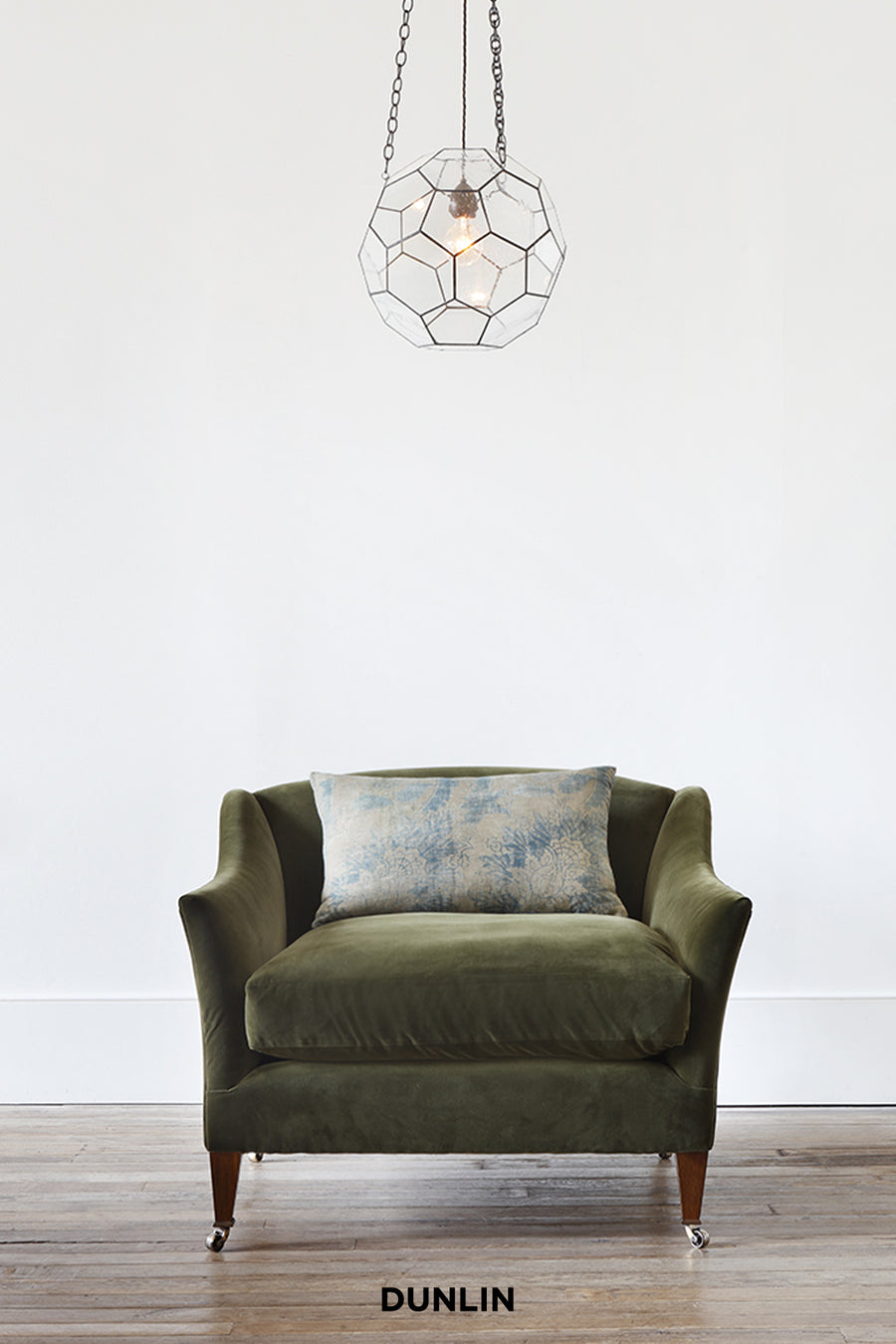 Green armchair with a patterned cushion under a geometric pendant light on a white wall.