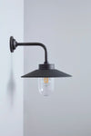 A modern wall-mounted sconce with a matte black metal arm and conical shade, featuring a clear glass globe bulb with a visible warm filament, mounted on a light gray wall.